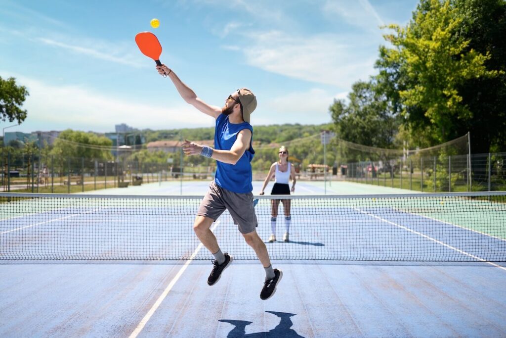 A player jumps to hit an overhead shot during a pickleball game on an outdoor court, while their partner watches from the opposite side of the net.
